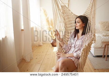 Beautiful Young Dark Haired Female In Trendy Striped Nightwear Relaxing In Swing. Attractive Brunett