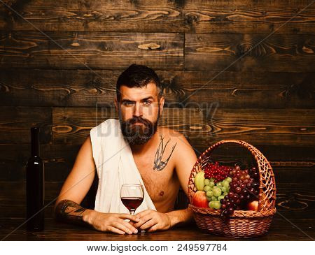 Man With Beard Holds Glass Of Wine On Vintage Wooden Background. God Bacchus With Serious Face Weari