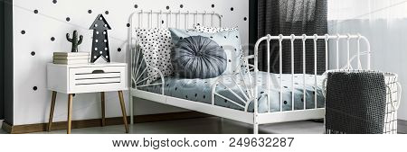 Wooden Bedside Table With Books And Lamp Standing By The White Metal Bed With Dotted Sheets In Brigh