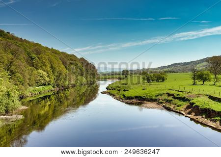 A View Of The River Lune Near Lancaster On A Sunny Day, With Green Fields And Wooded Slopes.