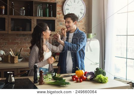 Happy Couple Cooking Healthy Food Together, Eating Cucumber, Feeding Each Other In Their Loft Kitche