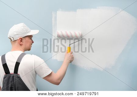 A Man Working In White T-shirt And Cap And Overalls Paints The Wall With A White Paint Roller.