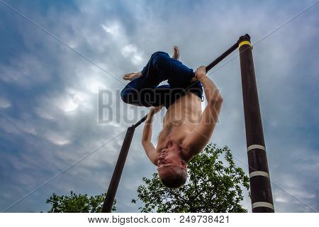 A Man With A Bare Torso In Blue Pants Upside Down Doing His Morning Exercises On A Horizontal Bar Ag
