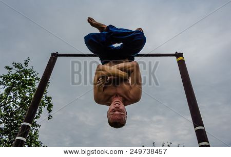 A Man With A Bare Torso In Blue Pants Upside Down Doing His Morning Exercises On A Horizontal Bar Ag