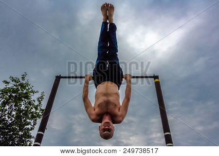 A Man With A Bare Torso In Blue Pants Upside Down Doing His Morning Exercises On A Horizontal Bar Ag