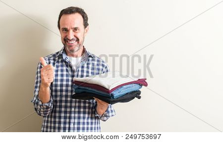 Senior man holding folded laundry clothes happy with big smile doing ok sign, thumb up with fingers, excellent sign