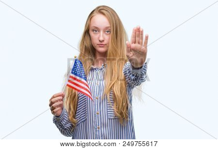 Blonde teenager woman holding flag of United States of America with open hand doing stop sign with serious and confident expression, defense gesture