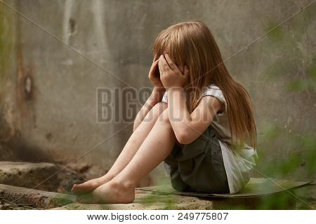 Portrait Of Crying Blond Girl Orphan With Long Matted Hair And Holen Knees Under The Dirty City Wall