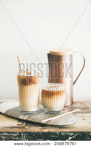 Iced Coffee In Tall Glasses With Milk And Straws On Board Over Rustic Wooden Table, White Wall And J