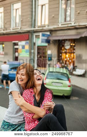 Two Young Girlfriends Hugging, Laughting And Sitting Near Road. One Girl With Long Brunette Hair In 
