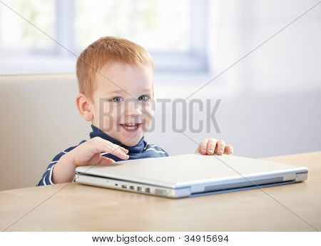 Adorable niño sentado en la mesa, sonriendo con ordenador portátil.