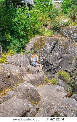 Young Woman Sitting On Rocks Around Dramatic And Rugged Mclaren Falls, Tauranga New Zealand.