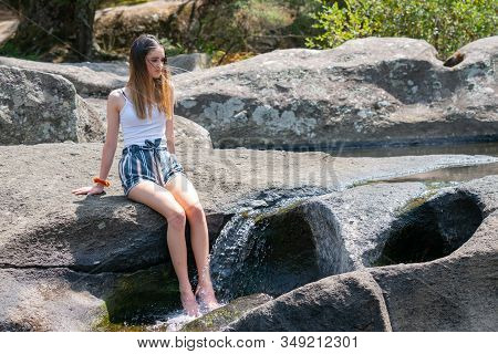 Attractive Teenage Girl Sitting On Rock Ledge With Legs Splashing Cool Water From Small Rock Waterfa