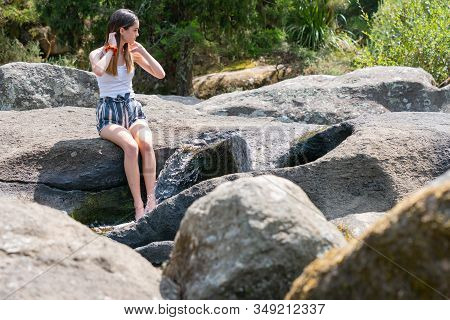 Attractive Teenage Girl Sitting On Rock Ledge With Legs Splashing Cool Water From Small Rock Waterfa