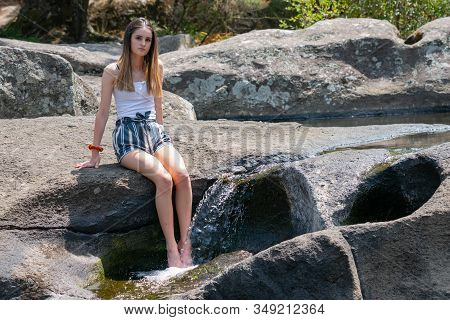 Attractive Teenage Girl Sitting On Rock Ledge With Legs Splashing Cool Water From Small Rock Waterfa