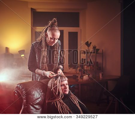 Attractive Dreadlock Master Is Working On Dreads For Pretty Blond Girl In The Dark Room.