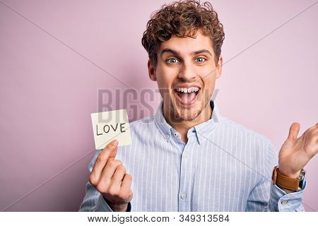 Young blond romantic man with curly hair holdiing paper with love message for valentines day very happy and excited, winner expression celebrating victory screaming with big smile and raised hands