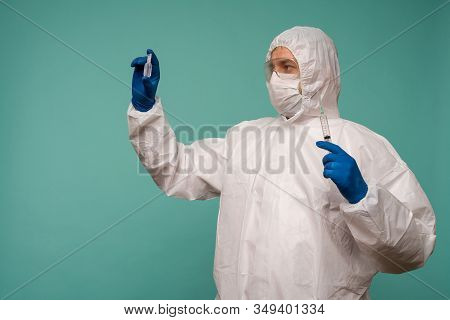 A Male Doctor In Protective Overalls And A Mask Holds An Ampoule With A Vaccine And Syringe In His H