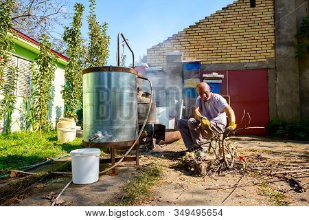 Man Throws Dry Branches Into The Firebox Of A Homemade Distillery Making Moonshine Schnapps, Alcohol