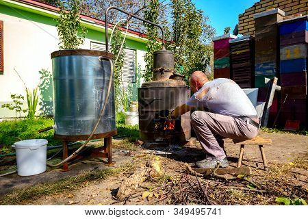 Man Throws Dry Branches Into The Firebox Of A Homemade Distillery Making Moonshine Schnapps, Alcohol