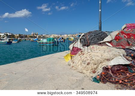 Fishing Nets Stand On A Concrete Jetty In Marsaxlokk, Malta. Typical Luzzu Fishing Boats Can Be Seen