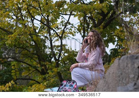 Young Pretty Girl Is Talking On The Phone While Sitting On A Large Stone At Autumn Background