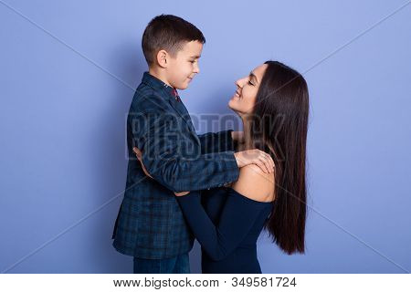 Image Of Positive Sweet Little Boy Wearing Dark Blue Suit, Looking At His Mother, Being On Good Term