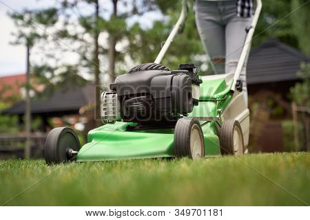 Close Up Of Female Legs Of Gardener Using Green Lawn Mower On Backyard. Selective Focus Of Woman Wor