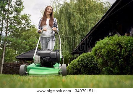 Front Below View Of Pretty Young Smiling Woman Using Lawn Mower On Backyard. Female Gardener With Lo