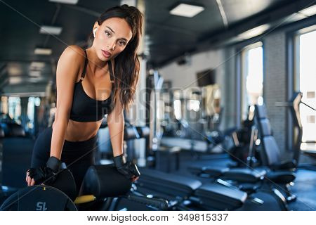Front View Of Young Woman Using Wireless Headpohnes, Doing Hyperextension At Gym, Looking At Camera.