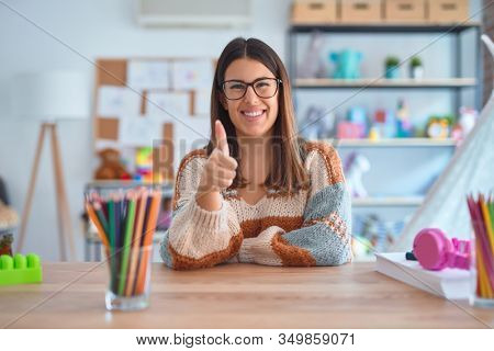 Young beautiful teacher woman wearing sweater and glasses sitting on desk at kindergarten doing happy thumbs up gesture with hand. Approving expression looking at the camera showing success.