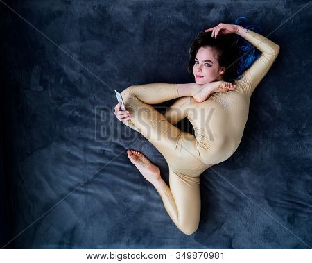 A Female Acrobat Lies On A Gray Bedspread In A Curved Pose And Uses A Mobile. A Woman Contortionist 