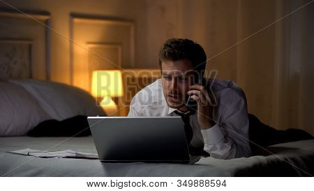Man In Suit Lying On Bed Working On Laptop And Talking On Phone, Busy Lifestyle