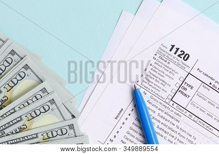 1120 Tax Form Lies Near Hundred Dollar Bills And Blue Pen On A Light Blue Background. Us Corporation