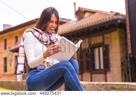 A Young Latin Woman With A White T-shirt, Jeans And A Scarf Enjoying Reading A Book, In The Old Town