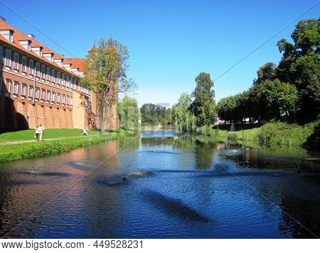 In Summer, People Walk Along The Bank Of A Moat With Water Around A Medieval Brick Castle With Tower