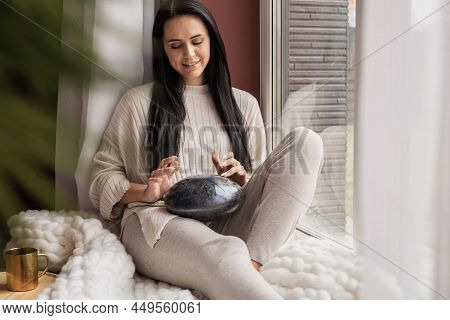 Smiling Young Woman Stroking Playing Glucophone Metal Tongue Drum Sitting On Windowsill