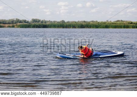Caucasian Little Female Kid Sitting On Sup Board On Small Lake Trying To Stand Up On Feet In Life Ja