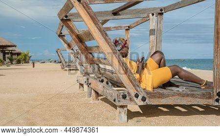 Africa Woman With African Colorful Headdress Sitting On A Bench In The Village Of Keta With Cellphon