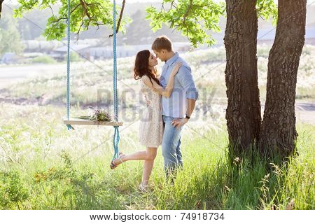 Young kissing couple under big tree with swing at sunset