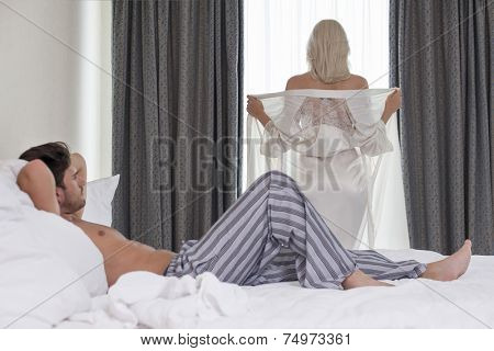 Full length of young man looking at woman removing robe at window