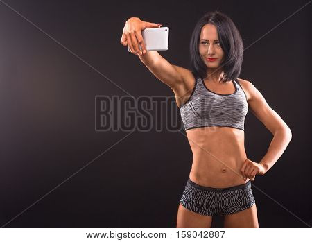 Sports, fitness, bodybuilding concepts. Bodybuilder woman posing with her hand on hip and holding mobile or smart phone in front of her. Pretty fitness lady making self photos.