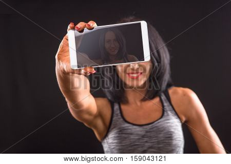 Sports or athletic lady holding mobile or smart phone in front of her and smiling for camera while posing isolated on black background in studio.