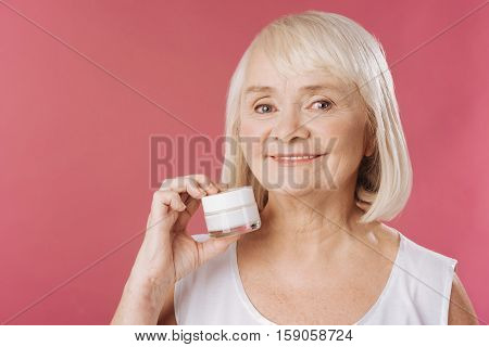 Fighting with wrinkles. Positive elderly grey haired woman standing against pink background and holding a bottle of cream while fighting with the age