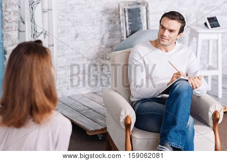 Positive atmosphere. Cheerful handsome professonal psychotherapst smiling and making notes while talkign with his patient