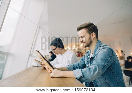 Men are sitting in front of the table near window. Happy young guy is loooking at menu and making order in restaurant