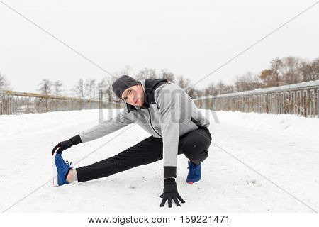 fitness, sport, people, exercising and healthy lifestyle concept - young man stretching leg and warmig up on snow covered winter bridge