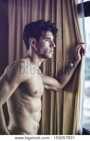 Sexy handsome young man standing shirtless in his bedroom next to window curtains