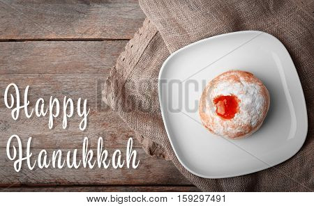 Plate with tasty doughnut on wooden background. Hanukkah celebration concept. Text HAPPY HANUKKAH