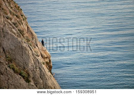 On a rock in the sea is a man and a fisherman is fishing on the fishing rod. The Rock is in the sea. Seascape on a sunny day. Sunset. On the rock plants grow. On the sea is calm. Extreme hobby.